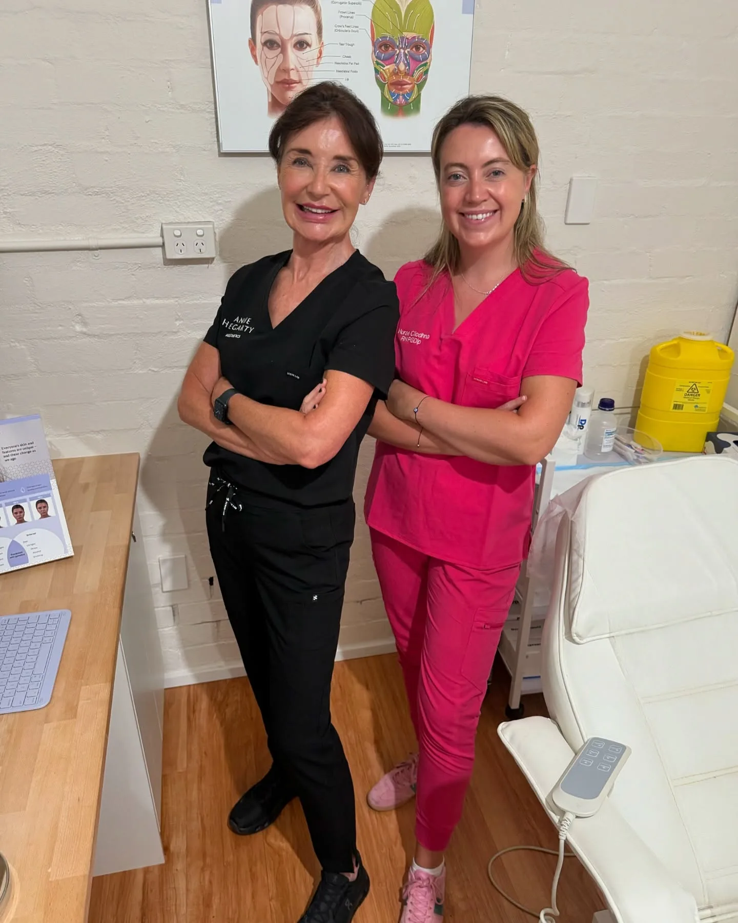 Two practitioners in the treatment room: Nurse Cliodhna in pink scrub embroidered 'Nurse Cliodhna RN.PGDip', alongside an in-clinic associate, both in the white-brick room